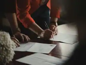 Pessoa a assinar um contrato com caneta sobre uma mesa de madeira com documentos.