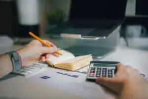 Pessoa a usar uma calculadora enquanto escreve num caderno com gráficos financeiros na mesa.