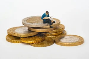 Figura em miniatura sentada sobre moedas enquanto lê, representando poupança ou investimento.